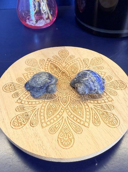 Two Blue and grey crystal turtles on a decorative wooden coaster in front of a vase and black jar