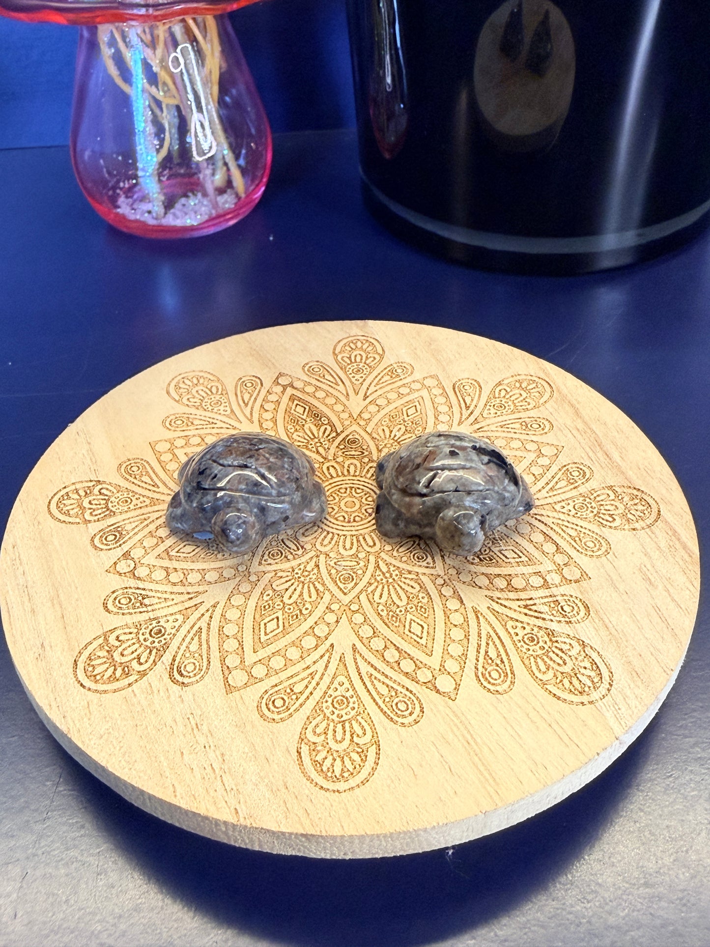 Two grey speckled crystal turtles on a decorative wooden coaster in front of a vase and black jar