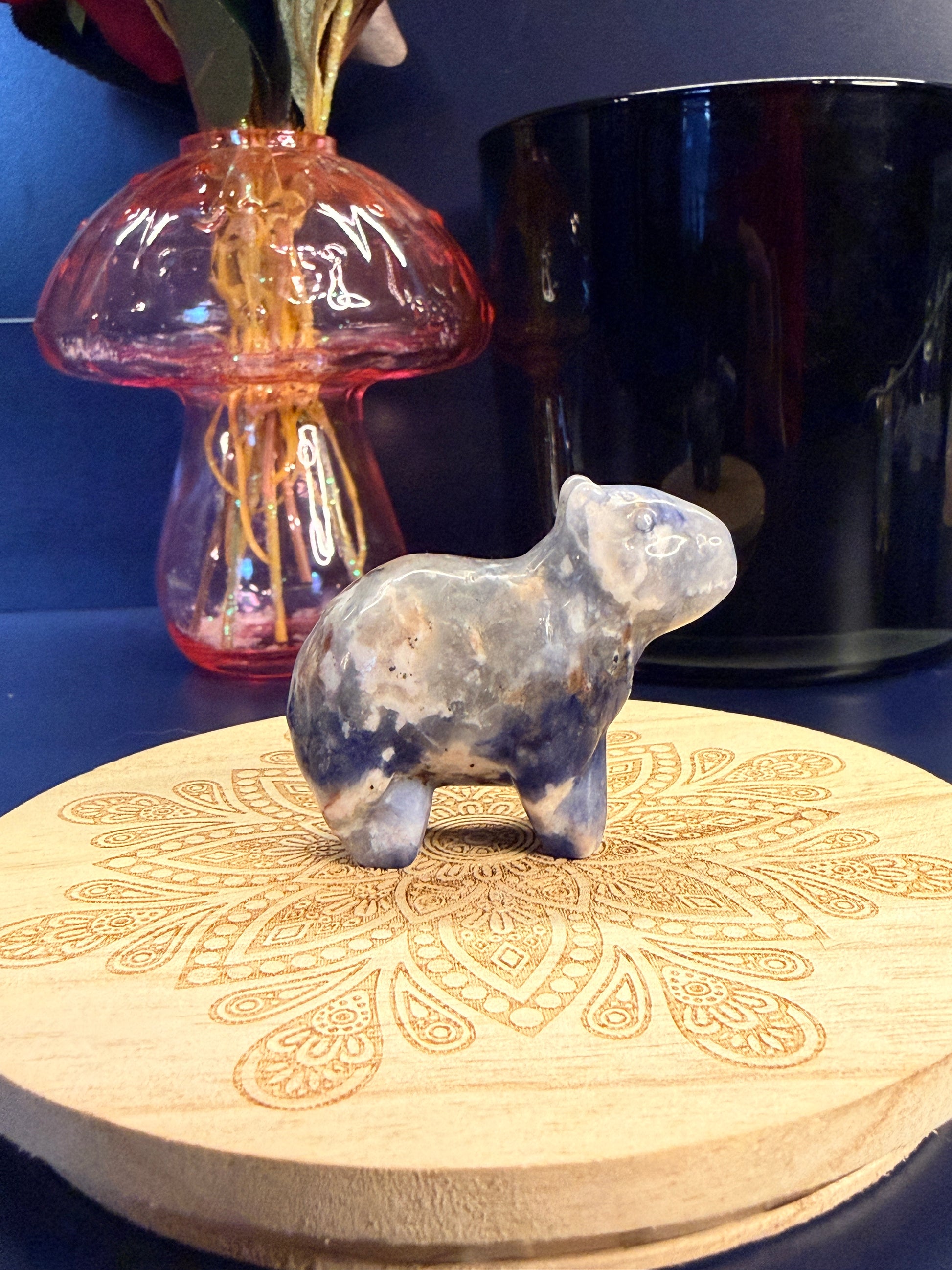 A sodalite capybara crystal sitting on a decorative wooden coaster in front of a glass mushroom and black jar