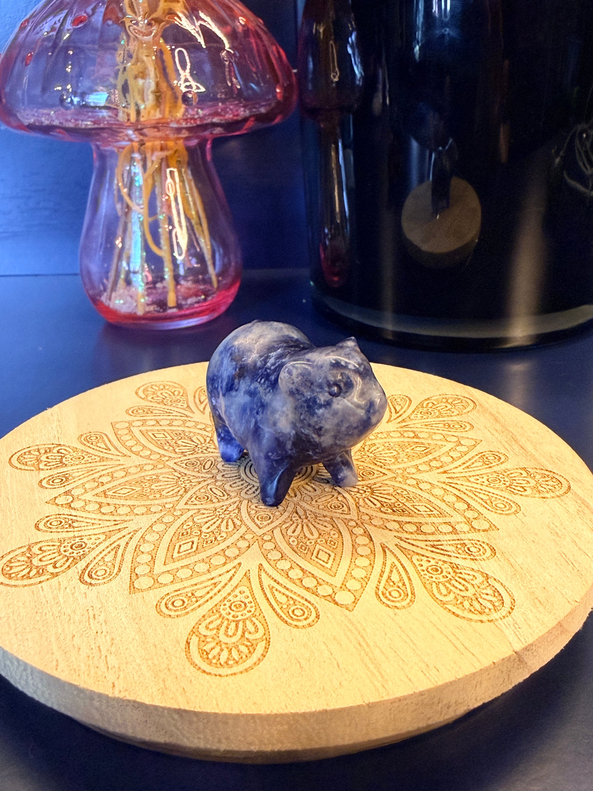 A sodalite pig crystal sitting on a decorative wooden coaster in front of a glass mushroom and black jar