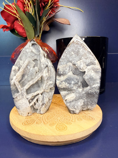 Two sparkly gray sphalerite freeforms on a wooden coaster in front of a red flower in a vase and black jar