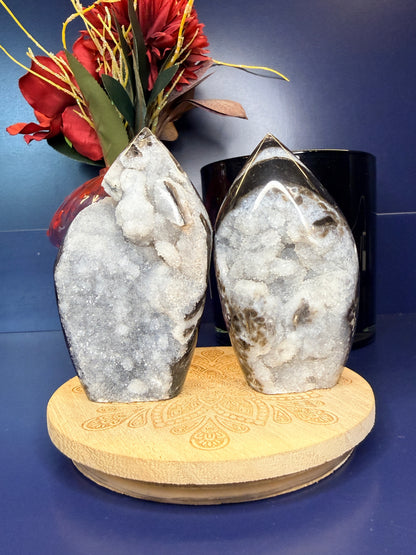 Two sparkly gray sphalerite freeforms on a wooden coaster in front of a red flower in a vase and black jar