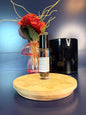 Body Oil Vial sitting on a decorative wooden coaster in front of a red flower and black jar