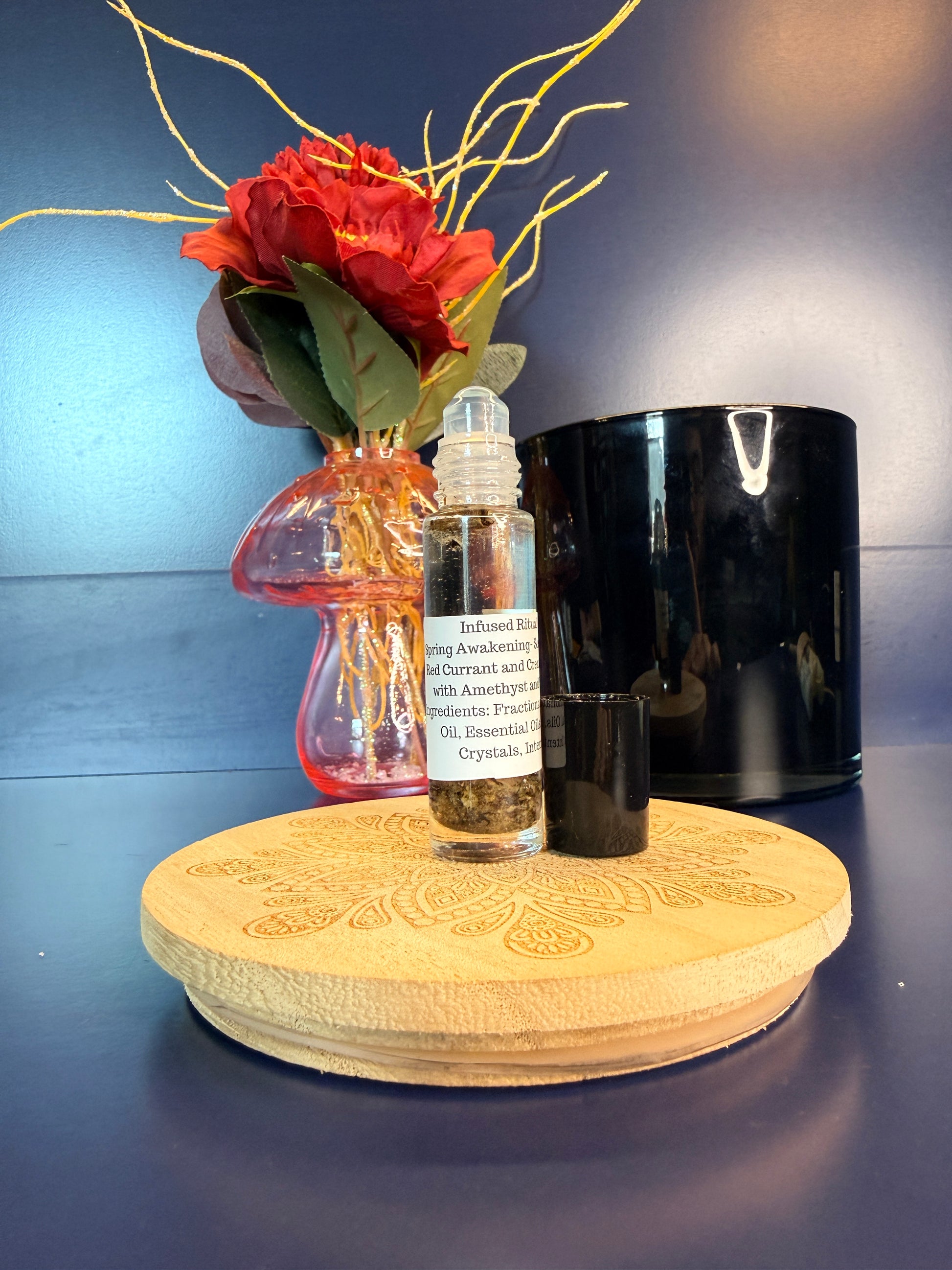 Body Oil Vial sitting on a decorative wooden coaster in front of a red flower and black jar