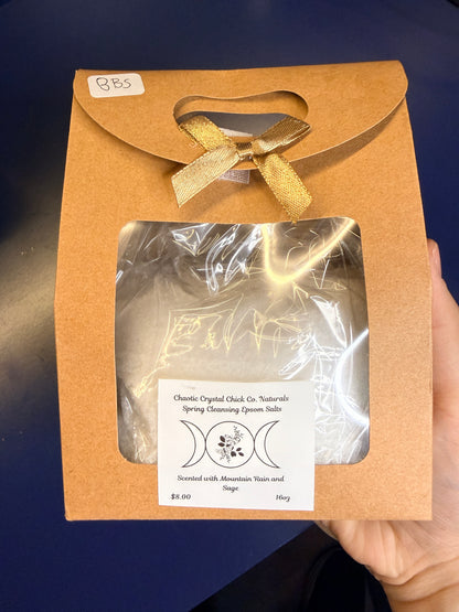 Hand holding white epsom salt in a brown paper container with a bow on a blue background