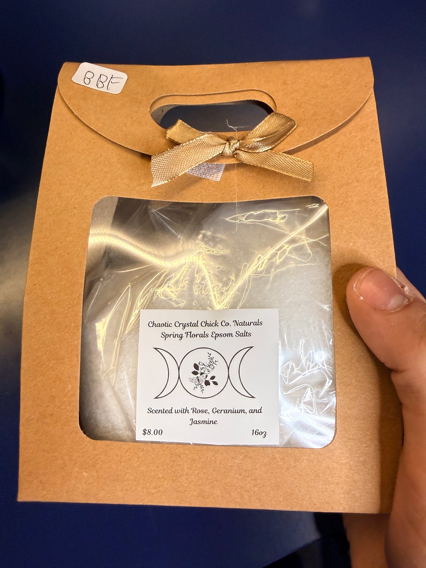 Hand holding white epsom salt in a brown paper container with a bow on a blue background
