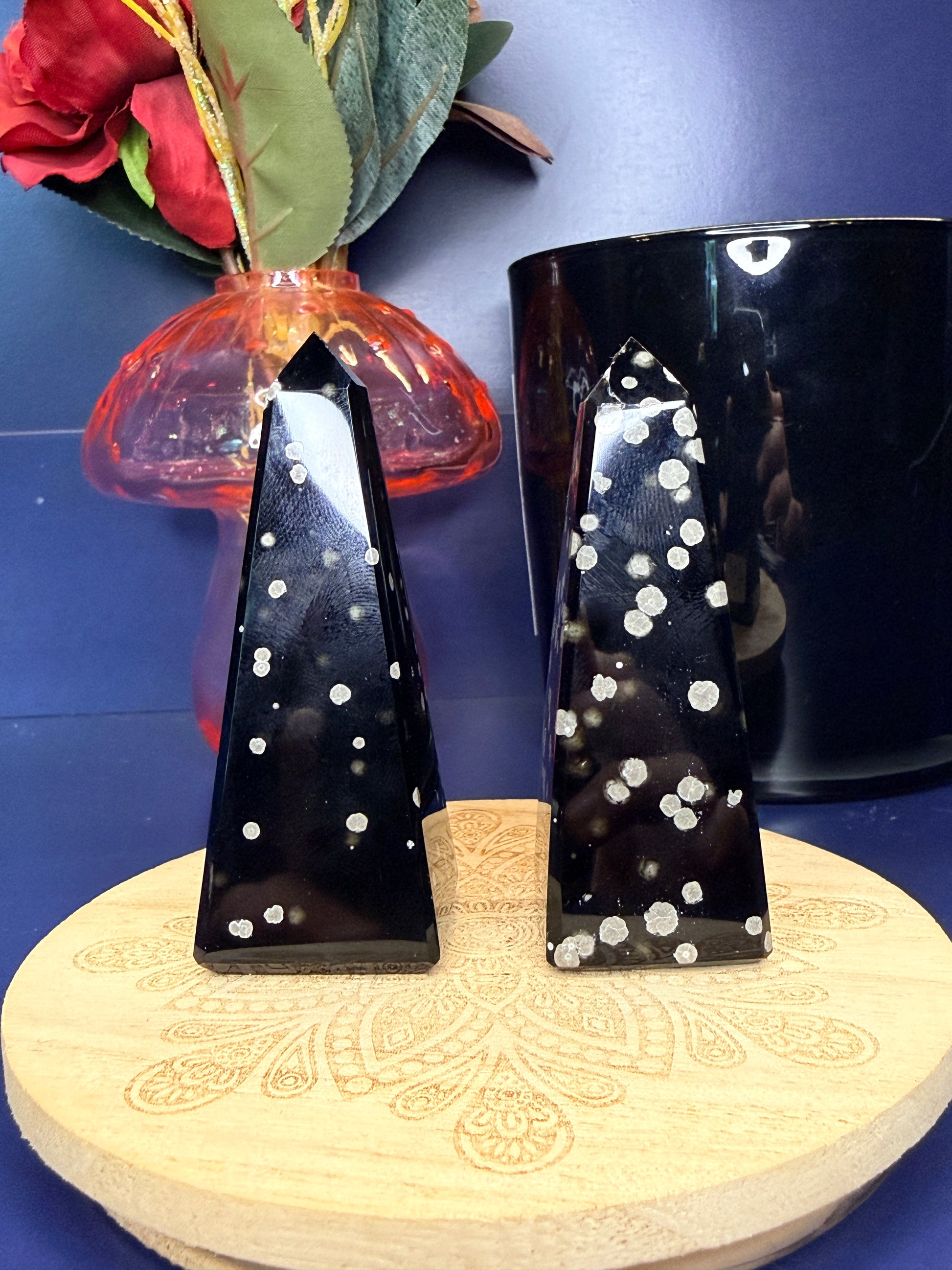 Two black and grey speckled stardust obsidian towers on a wooden coaster in front of a red flower in a vase and black jar