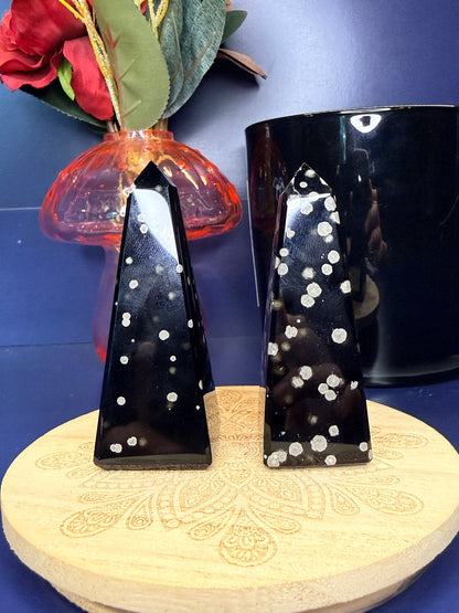 Two black and grey speckled stardust obsidian towers on a wooden coaster in front of a red flower in a vase and black jar