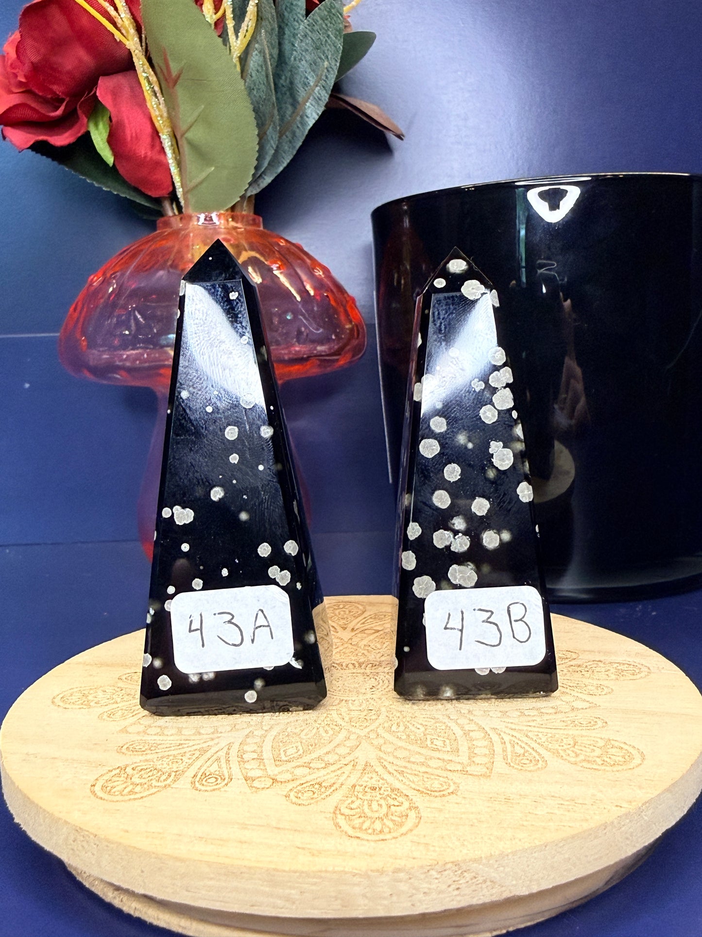 Two black and grey speckled stardust obsidian towers on a wooden coaster in front of a red flower in a vase and black jar