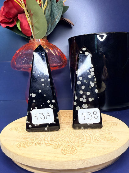 Two black and grey speckled stardust obsidian towers on a wooden coaster in front of a red flower in a vase and black jar