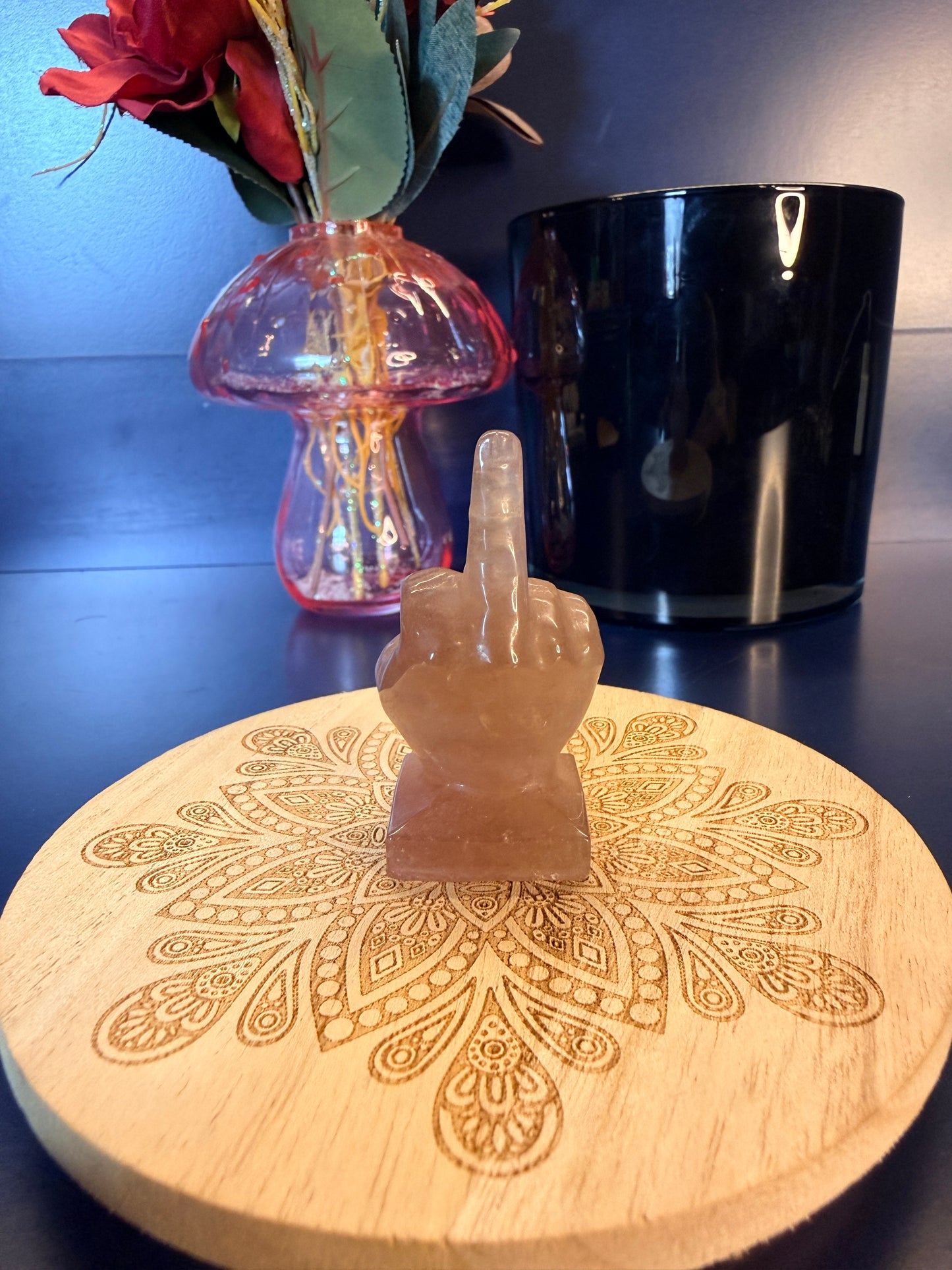 A pink crystal middle finger sitting on a decorative wooden coaster in front of a glass mushroom and black jar