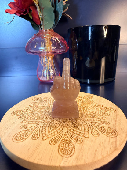 A pink crystal middle finger sitting on a decorative wooden coaster in front of a glass mushroom and black jar
