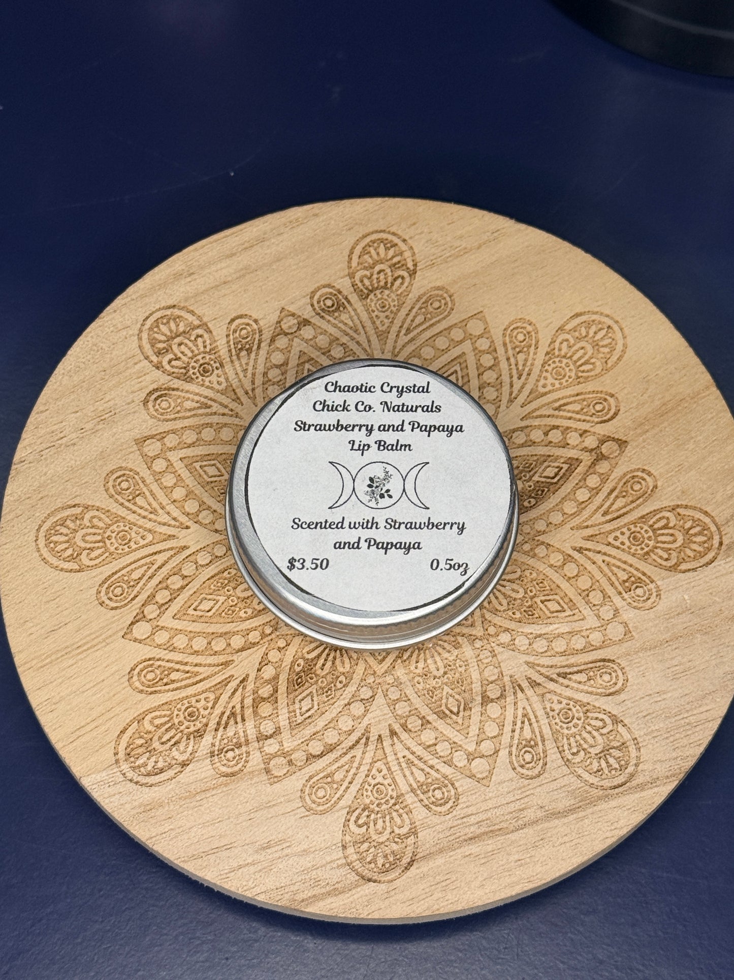 Circular labeled lip balm container on a decorative wooden coaster on a blue background