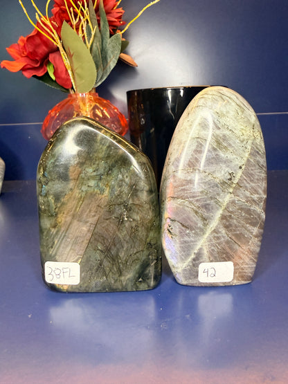 Two large grey labradorite free forms in front of a red flower and black jar on a blue background