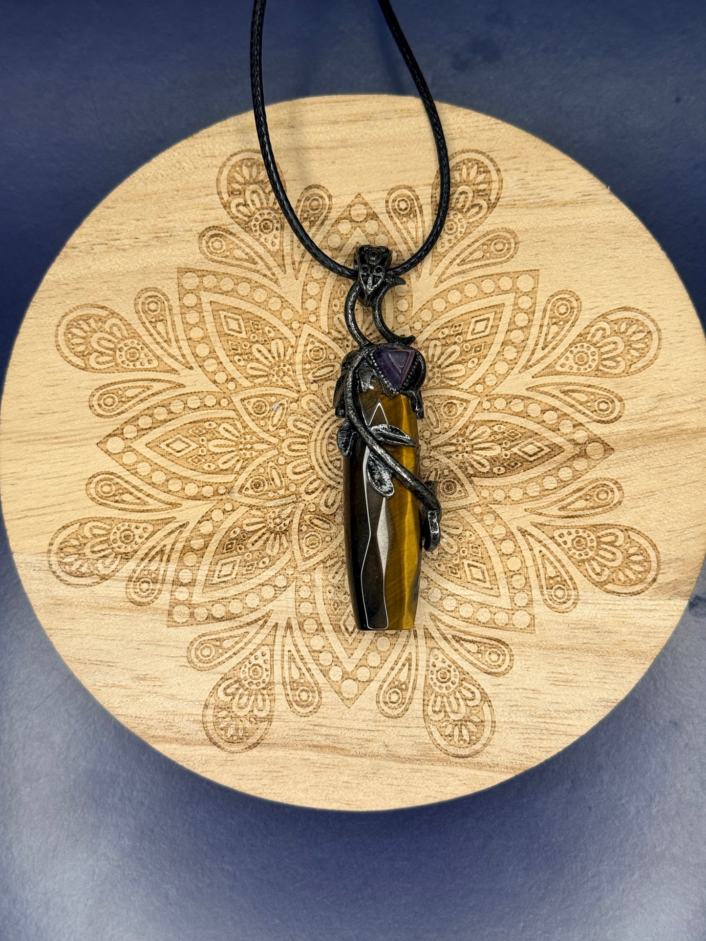 Intricate tiger's eye pendant with leaves wrapping around it sitting on a decorative wooden coaster on a blue background