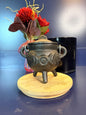 A cast iron cauldron with a triple moon design sitting on a decorative wooden coaster in front of a red flower and black jar