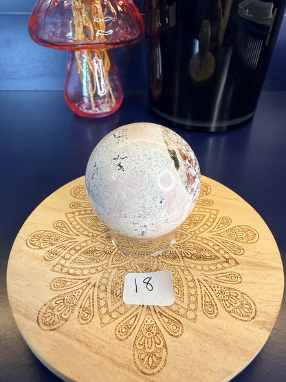 White and magenta unicorn jasper sphere on a decorative wooden coaster in front of a mushroom vase and black jar