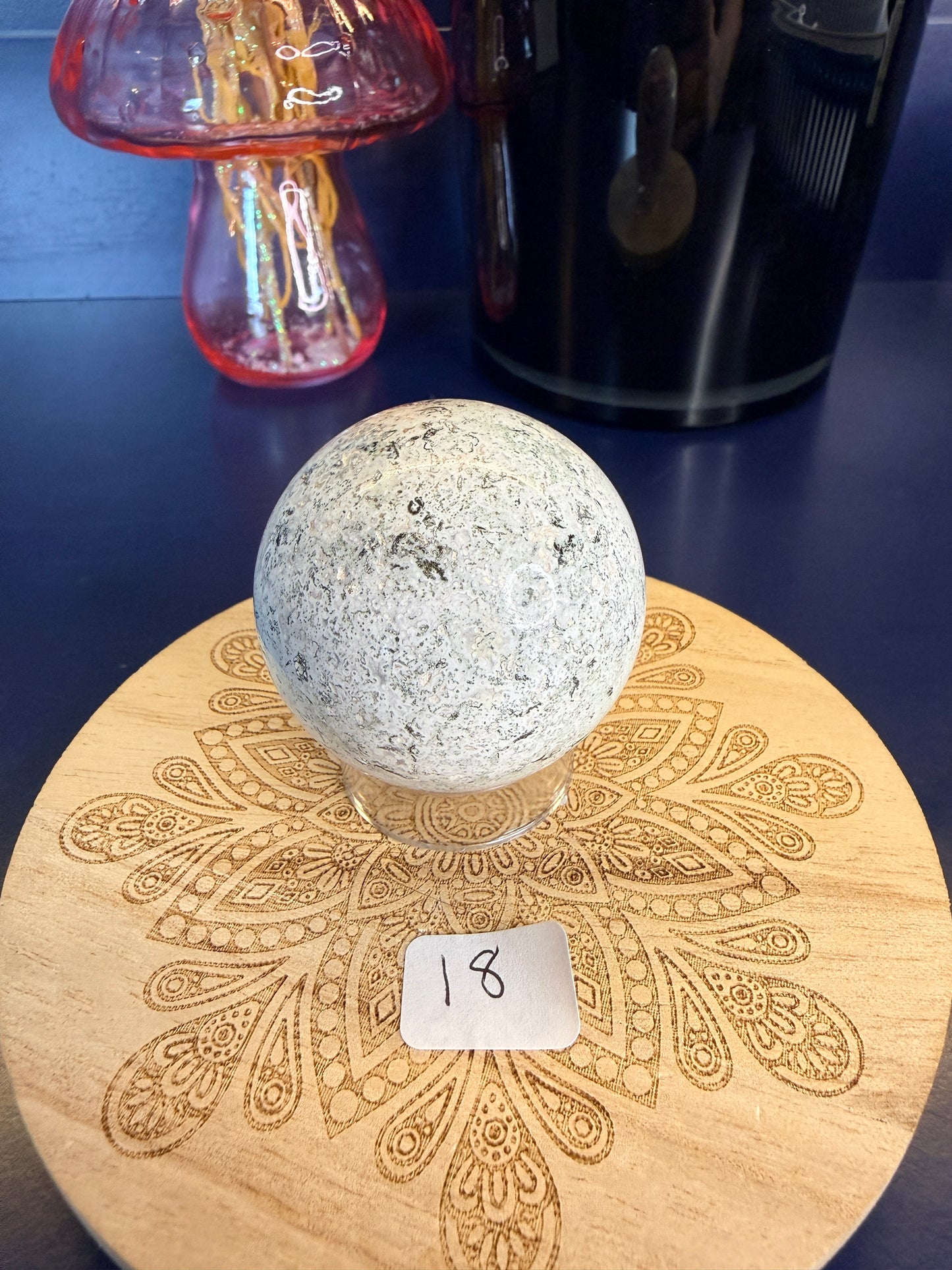 White speckled unicorn jasper sphere on a decorative wooden coaster in front of a mushroom vase and black jar