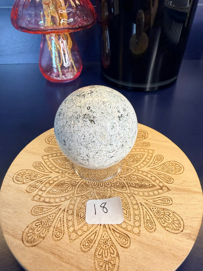 White speckled unicorn jasper sphere on a decorative wooden coaster in front of a mushroom vase and black jar