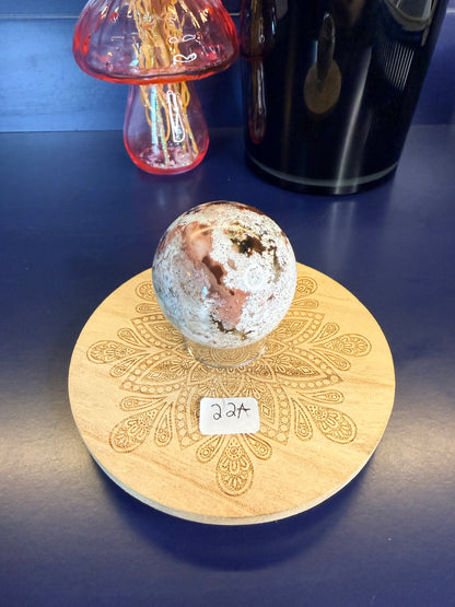 White and magenta unicorn jasper sphere on a decorative wooden coaster in front of a mushroom vase and black jar