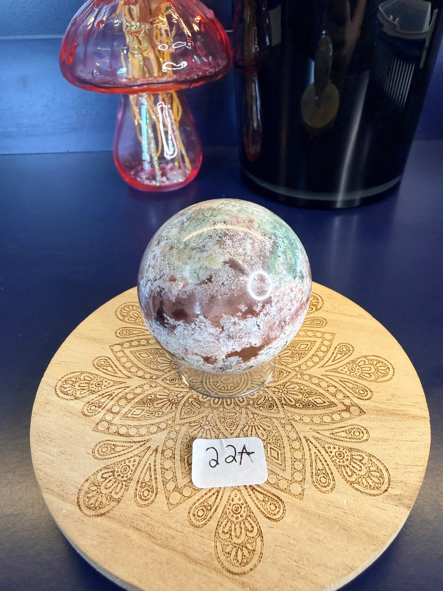 White and magenta unicorn jasper sphere on a decorative wooden coaster in front of a mushroom vase and black jar