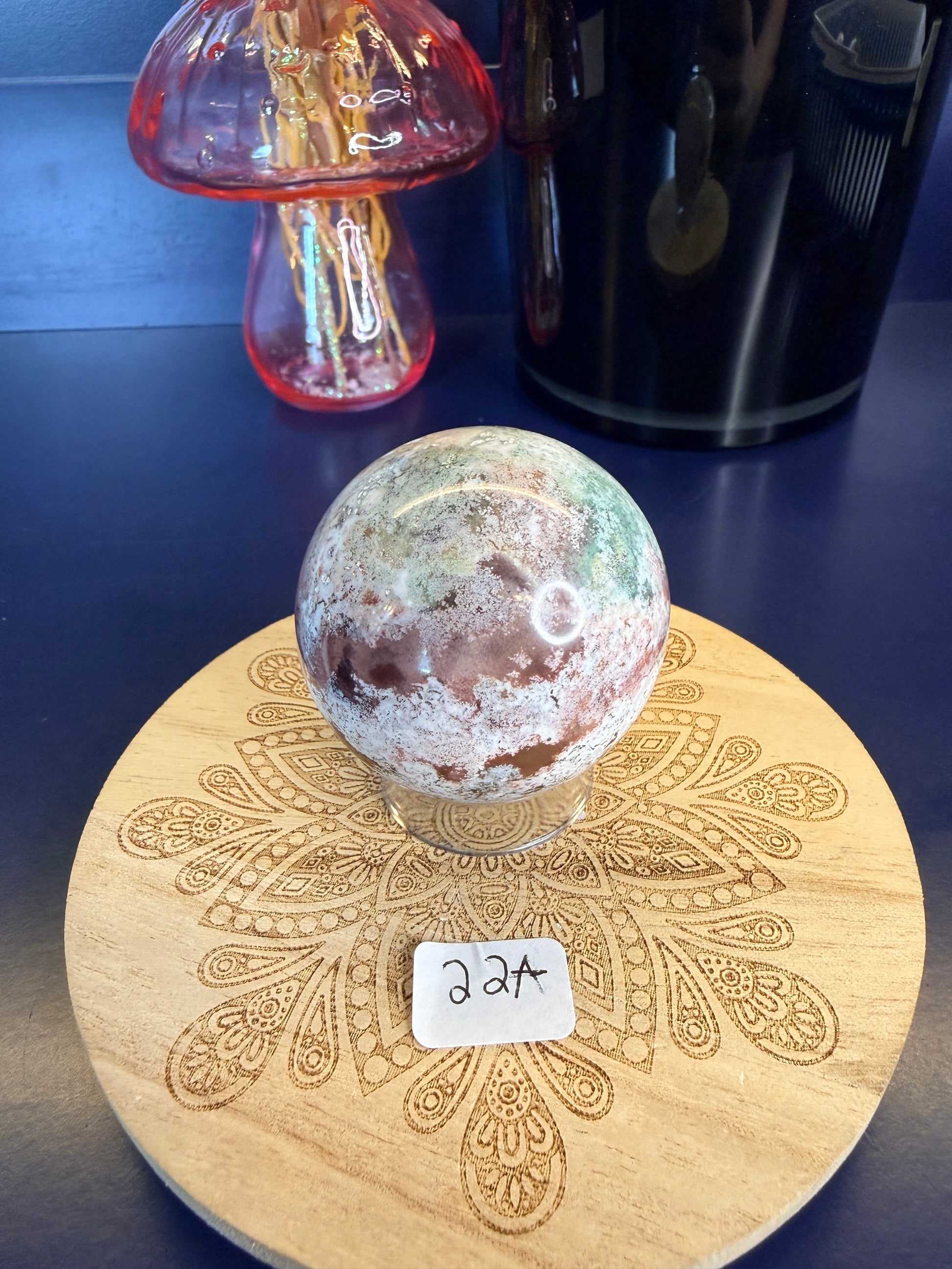 White and magenta unicorn jasper sphere on a decorative wooden coaster in front of a mushroom vase and black jar