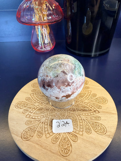 White and magenta unicorn jasper sphere on a decorative wooden coaster in front of a mushroom vase and black jar