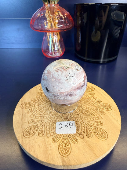 White and magenta unicorn jasper sphere on a decorative wooden coaster in front of a mushroom vase and black jar