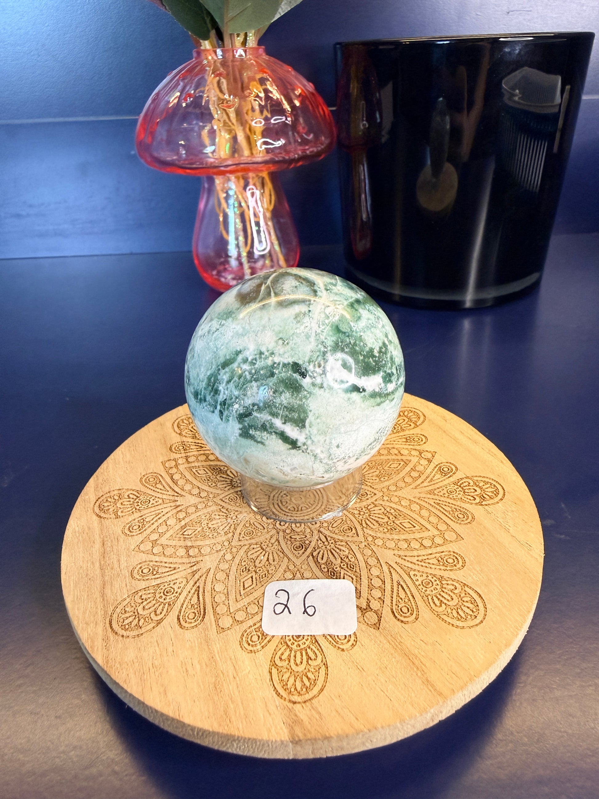 White and green unicorn jasper sphere on a decorative wooden coaster in front of a mushroom vase and black jar
