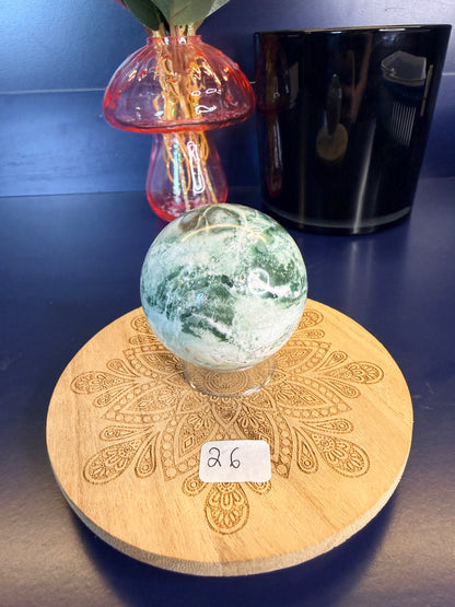 White and green unicorn jasper sphere on a decorative wooden coaster in front of a mushroom vase and black jar