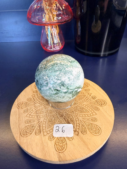 White and green unicorn jasper sphere on a decorative wooden coaster in front of a mushroom vase and black jar