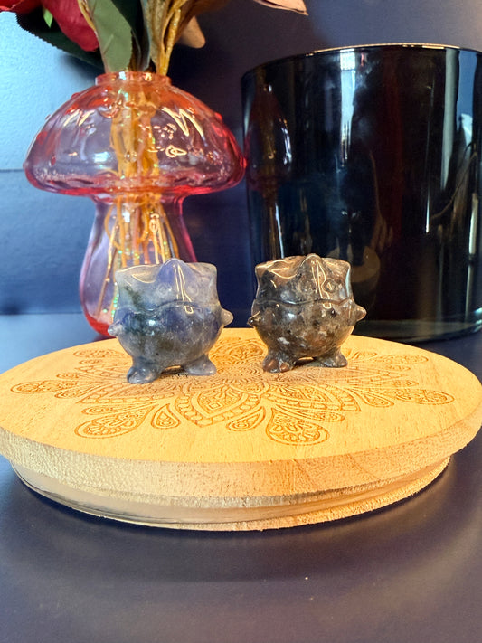 Two Togepi-inspired shapes, one sodalite and one yooperlite, on a wooden coaster in front of a glass mushroom and black jar