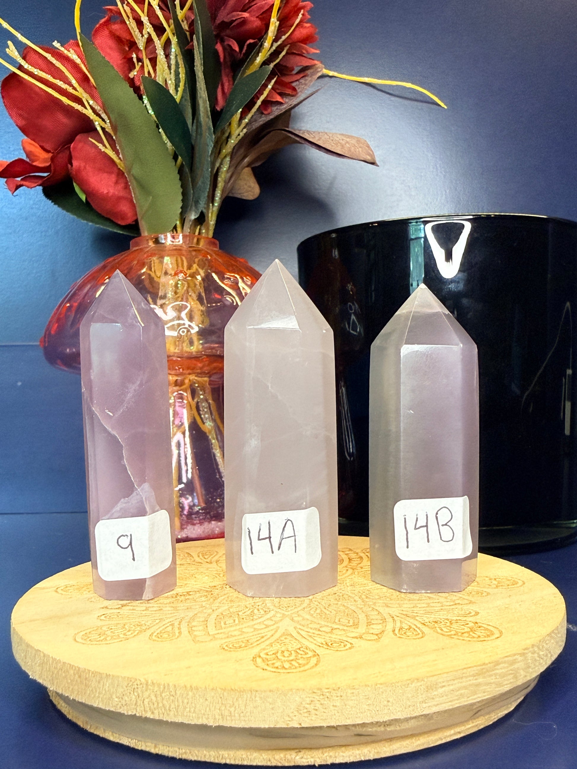 Three lavender yttrium fluorite towers on a wooden coaster in front of a red flower in a vase and a black jar
