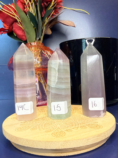 Three lavender and green yttrium fluorite towers on a wooden coaster in front of a red flower in a vase and a black jar