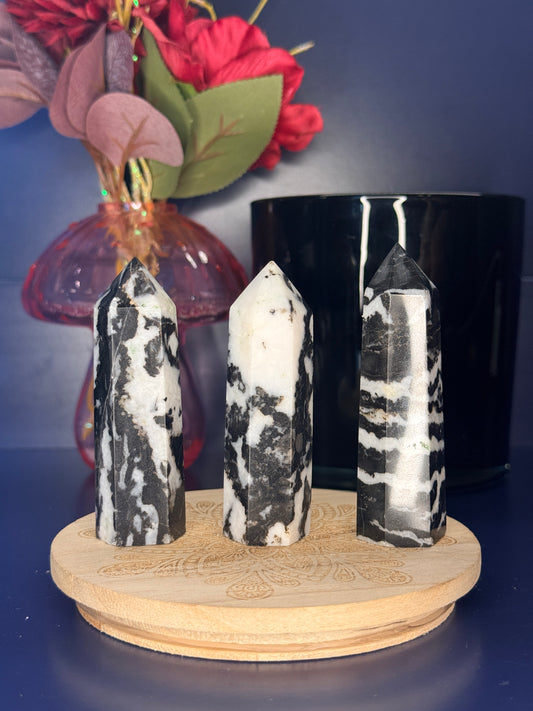 Three black and white zebra jasper towers in front of a pink vase and black jar on a wooden coaster against a blue background
