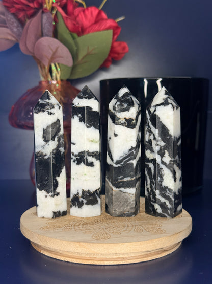 Four black and white zebra jasper towers in front of a pink vase and black jar on a wooden coaster against a blue background