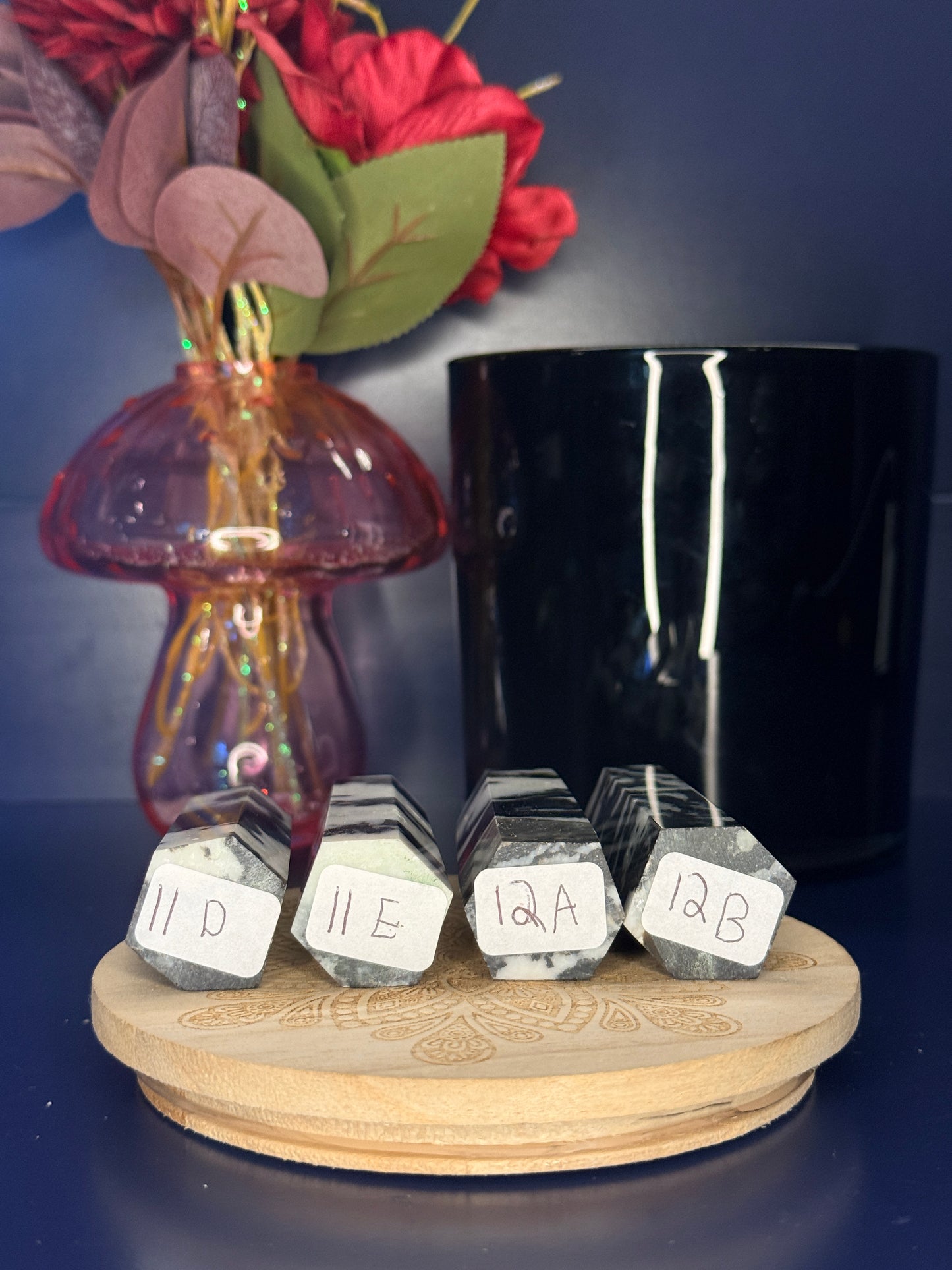 Four black and white zebra jasper towers with labels in front of a pink vase and black jar on a wooden coaster against a blue background