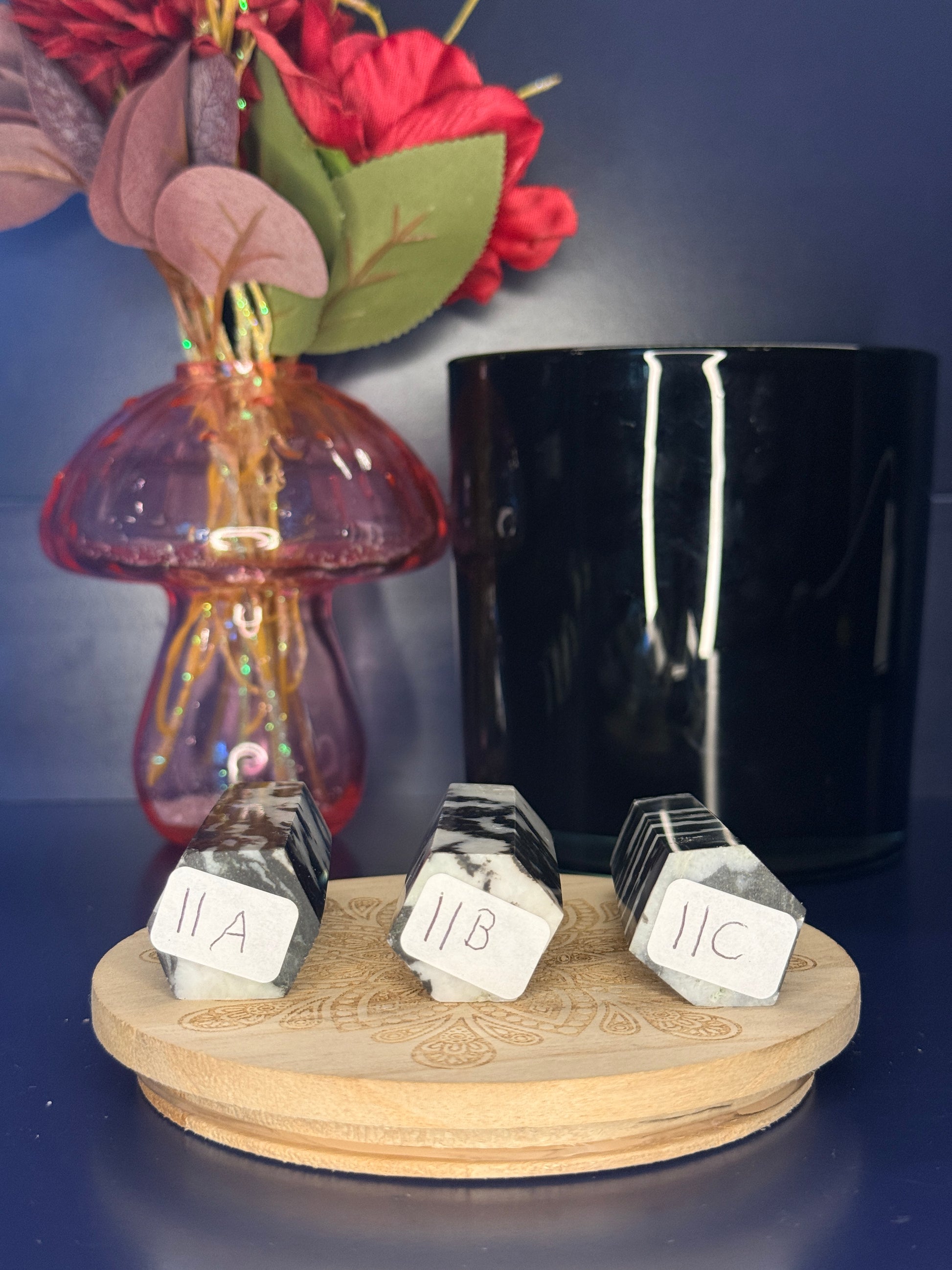 Three black and white zebra jasper towers with labels in front of a pink vase and black jar on a wooden coaster against a blue background